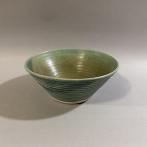 May include: A handmade, green ceramic bowl with a textured, ridged exterior. The bowl has a wide, open top and a slightly off-white base. The interior has a smooth, glossy finish. The bowl is approximately 6 inches in diameter.
