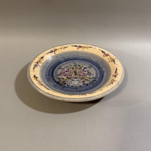 May include: A decorative ceramic plate with a blue and cream color scheme. The center features a blue spiral design with two pink birds and floral accents. The rim has a cream border with floral and leaf patterns. The plate is round and has a slightly raised edge.