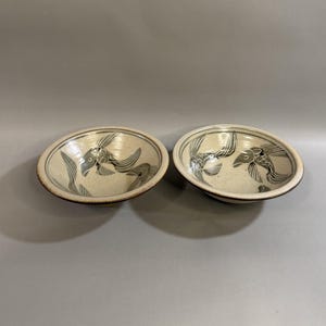 May include: Two ceramic bowls with a beige background and a dark gray fish design. The bowls are oval-shaped with a dark brown rim. The fish design is repeated on both bowls, creating a matching set. The bowls are likely handmade.