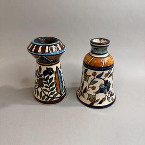May include: Two ceramic candle holders with hand-painted floral patterns. The left holder has a wide, flared top with brown and white stripes, while the right holder features a narrower neck and a bell-shaped base with blue, orange, and green motifs.