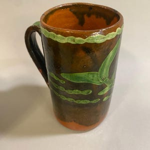Puede incluir: Taza de cer&aacute;mica hecha a mano con base marr&oacute;n y exterior marr&oacute;n oscuro. El interior es naranja y el borde est&aacute; decorado con pintura verde. Dise&ntilde;os verdes en forma de hoja adornan el exterior de la taza. El asa es negra.