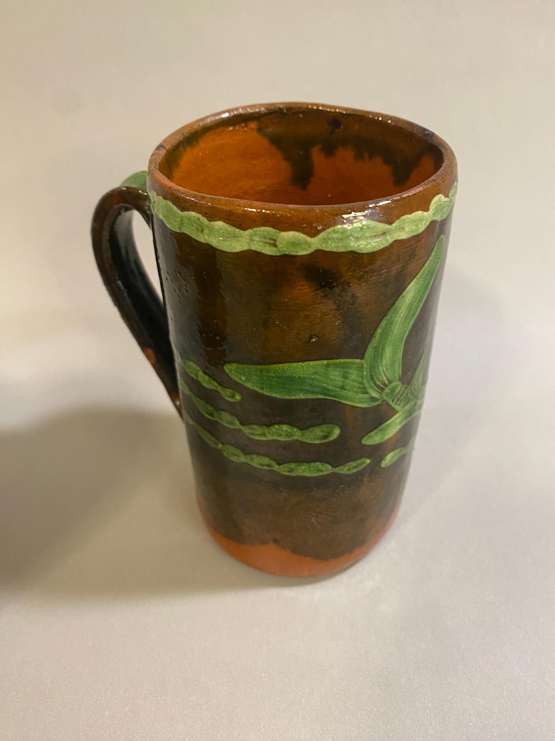 Puede incluir: Taza de cer&aacute;mica hecha a mano con base marr&oacute;n y exterior marr&oacute;n oscuro. El interior es naranja y el borde est&aacute; decorado con pintura verde. Dise&ntilde;os verdes en forma de hoja adornan el exterior de la taza. El asa es negra.