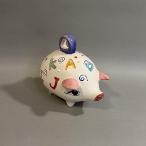 May include: A white ceramic piggy bank with colorful letters and a blue handle. The piggy bank has a pink snout and ears, and a black eye with eyelashes. The letters are in various colors, including red, yellow, and green.