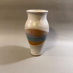 May include: A handmade ceramic vase with a white base and a colorful band of orange, blue, and yellow. The vase has a slightly flared rim and a smooth, glossy finish. The vase is approximately 8 inches tall.