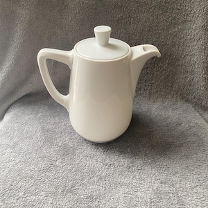 May include: A white ceramic teapot with a lid and a spout. The teapot is sitting on a gray fuzzy surface.