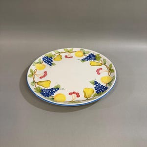 May include: White ceramic plate with a blue rim, decorated with a colorful fruit design. The design includes yellow pears, grapes, and red berries. A decorative plate for serving food.
