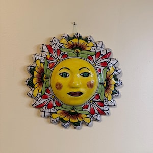 May include: A circular ceramic wall plaque featuring a central yellow sun face with green eyes, brown cheeks, and red lips. The sun is surrounded by a decorative border of yellow sunflowers, red petals, and a white grid pattern with blue dots.