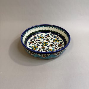 May include: A round ceramic bowl featuring a hand-painted floral pattern with yellow, blue, and green petals on a white background. The interior rim displays a blue and white triangular border, while the exterior features a scalloped pattern.