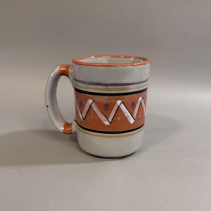 May include: A ceramic mug with a white base and a handle. The mug features a design of orange zigzags, black, yellow, and purple stripes. The rim and handle accents are orange.