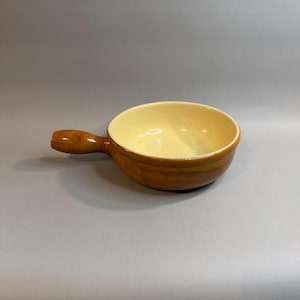 May include: A round ceramic bowl with a single handle extending from the side. The exterior features a glossy, warm brown glaze, while the interior is finished in a smooth, light cream color. The bowl has a wide, open top and a sturdy base.