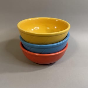 May include: A stack of three round ceramic bowls with a smooth, glossy finish. The top bowl is bright yellow, the middle bowl is light blue, and the bottom bowl is a vibrant orange. Each bowl features a subtle, repeating diagonal embossed pattern.