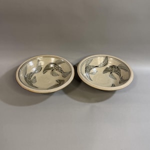 May include: Two ceramic bowls with a beige background and a dark gray fish design. The bowls are round with a slightly raised edge. The fish design is repeated in each bowl, with the fish swimming in different directions.