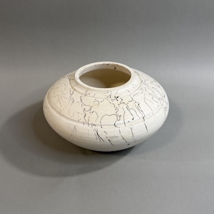 May include: A cream-colored ceramic vase with a wide, rounded body and a circular opening. The vase is decorated with black, abstract, linear patterns. The design is applied to the exterior surface of the vase.