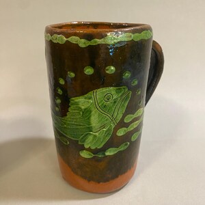 Puede incluir: Una taza de cer&aacute;mica hecha a mano con un esmalte marr&oacute;n, con un dise&ntilde;o de pez verde. La taza tiene un asa y est&aacute; decorada con burbujas verdes y un borde verde. La base es naranja.