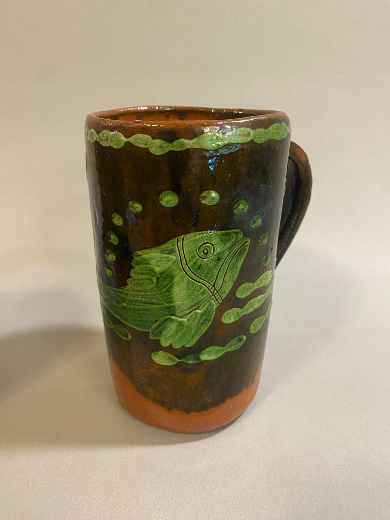 Puede incluir: Una taza de cer&aacute;mica hecha a mano con un esmalte marr&oacute;n, con un dise&ntilde;o de pez verde. La taza tiene un asa y est&aacute; decorada con burbujas verdes y un borde verde. La base es naranja.