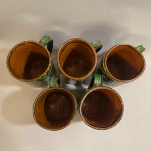 Puede incluir: Vista a&eacute;rea de cinco tazas de cer&aacute;mica hechas a mano. Las tazas tienen un exterior oscuro con asas verdes y un interior marr&oacute;n. Los bordes est&aacute;n decorados con un patr&oacute;n amarillo y verde. Las tazas est&aacute;n dispuestas en un patr&oacute;n circular.