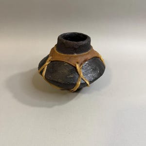 May include: A handcrafted, dark brown ceramic vase with a wide, rounded base and a narrow, cylindrical neck. The vase is adorned with a tan leather band secured with tan leather lacing. The overall design is rustic and earthy.