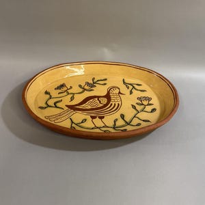 May include: Oval ceramic serving dish with a yellow interior and a brown rim. The dish features a brown bird design with floral accents. The bird is standing on a branch. The dish is likely handmade.