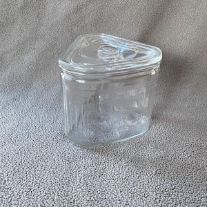May include: A clear glass jar with a triangular lid. The jar has a rectangular body with vertical lines etched into the glass. The lid has a small handle. The jar is empty.