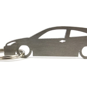 Może przedstawiać: Srebrny metalowy brelok do kluczy w kształcie samochodu Ford Focus. Brelok ma rozdzielony pierścień do mocowania kluczy.