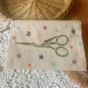 May include: A beige canvas zipper pouch featuring a green illustration of scissors and small colorful flowers. The pouch has a beige zipper and a fabric pull. The background includes a woven basket and a straw hat.