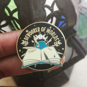May include: A black and gold enamel pin featuring the text "Devourer of Worlds" in a circle around a book with a quill pen and a crescent moon and stars.