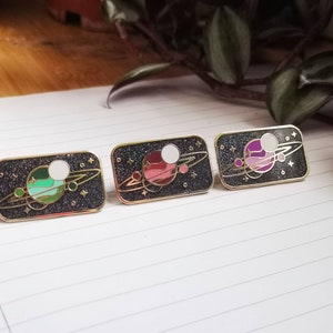May include: Three enamel pins with a black background and gold accents. Each pin features a white moon and a planet with a ring system. The planets are colored green, pink, and purple.
