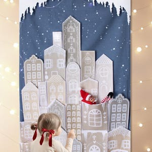 Peut inclure: Une enfant interagit avec une scène de village de Noël en papier. Le village présente des bâtiments beiges et blancs avec des détails complexes, sur fond bleu avec un effet neigeux. Une petite figurine d'elfe est nichée dans le village.