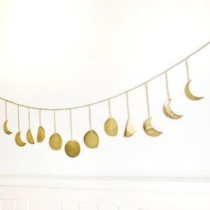 May include: A gold-coloured moon phase garland featuring various moon shapes suspended from a chain. The garland includes crescent moons, half moons, and full moons, creating a celestial theme. The metal has a hammered texture.