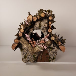 Peut inclure: Une petite maisonnette rustique pour oiseaux en pierre et en bois avec une ouverture de fenêtre. La maisonnette est décorée de fleurs roses et de branches.