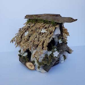 May include: A miniature fairy house made of wood and white stones, decorated with moss, dried flowers, and small white flowers. The house has a roof made of a piece of wood with a mossy texture.