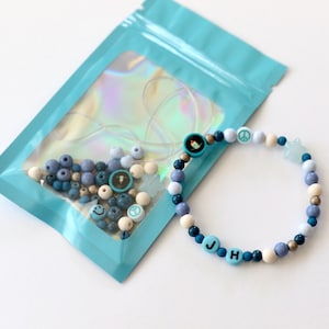 May include: A blue and white beaded bracelet with the letters "JH" and various charms, including peace signs, smiley faces, and stars. The bracelet is packaged in a blue resealable bag with a clear window.