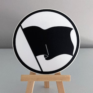May include: A black and white sticker with a black flag on a white background. The sticker is on a small wooden easel.