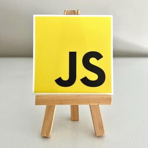 May include: A yellow square sticker with the letters "JS" in black, displayed on a small wooden easel.
