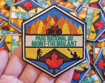 Könnte beinhalten: Ein sechseckiger gestickter Aufnäher mit blauem Hintergrund und weißem Rand. Der Aufnäher zeigt eine Bergkette mit einem roten Ahornblatt in der Mitte. Der Text "Parc National du Mont-Tremblant" ist in Weiß auf einem weißen Banner geschrieben. Der Aufnäher zeigt auch einen Mountainbiker und einen Kanuten.