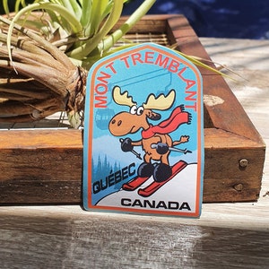 K&ouml;nnte beinhalten: Ein blauer und wei&szlig;er Aufkleber mit einem Cartoon-Elch, der Ski f&auml;hrt. Der Aufkleber zeigt "Mont Tremblant" in einem wei&szlig;en Bogen &uuml;ber dem Elch, "Quebec" in wei&szlig; unter dem Elch und "Canada" in wei&szlig; unten.
