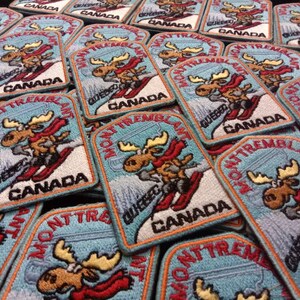 K&ouml;nnte beinhalten: Ein Stapel blauer und wei&szlig;er gestickter Aufn&auml;her mit einem Cartoon-Elch, der Ski f&auml;hrt, mit dem Text "Mont Tremblant" und "Quebec Canada".