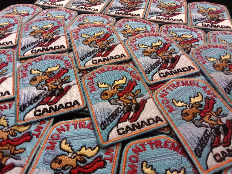 K&ouml;nnte beinhalten: Ein Stapel blauer und wei&szlig;er gestickter Aufn&auml;her mit einem Cartoon-Elch, der Ski f&auml;hrt, mit dem Text "Mont Tremblant" und "Quebec Canada".