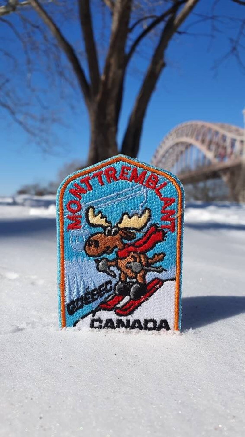 K&ouml;nnte beinhalten: Ein blauer und orangefarbener gestickter Aufn&auml;her mit einem Cartoon-Elch, der Ski f&auml;hrt. Der Aufn&auml;her tr&auml;gt die Aufschrift "Mont Tremblant Quebec Canada".
