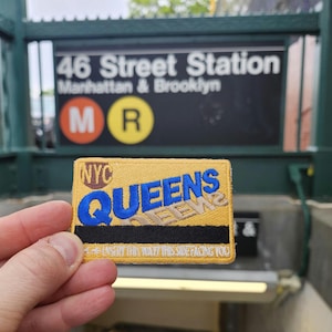 Può includere: Una toppa ricamata gialla e blu con la scritta "NYC QUEENS" e "INSERT THIS WAY/THIS SIDE FACING YOU". La toppa è tenuta davanti a un cartello della stazione della metropolitana che recita "46 Street Station Manhattan & Brooklyn".