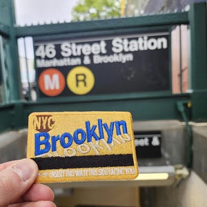 以下が含まれることがあります： 地下鉄の入り口の前で、青い文字で「Brooklyn」と書かれた黄色と黒のブルックリンのパッチが掲げられています。パッチには「Brooklyn」と「NYC」と書かれています。地下鉄の標識には「46 Street Station Manhattan & Brooklyn」と書かれています。