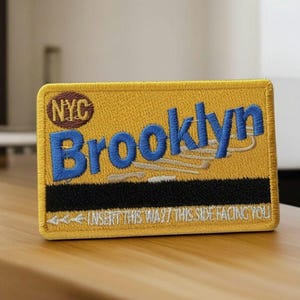 May include: Embroidered patch with "Brooklyn" in blue letters on a yellow background. The patch includes "NYC" in a brown circle and the text "INSERT THIS WAY/THIS SIDE FACING YOU" in white.