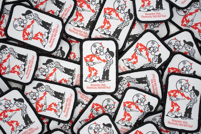 May include: A close-up of a pile of white fabric patches with black borders. Each patch features a cartoon illustration of a stick figure with a lightning bolt and the text "Remember Kids, Electricity Will Kill You!"