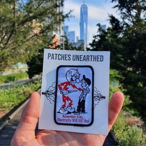 May include: A white patch with black trim featuring a cartoon illustration of a man in a red suit holding a knife and a boy in a blue shirt. The text on the patch reads "Remember Kids, Electricity Will Kill You!" The patch is held in a hand against a backdrop of a city skyline.
