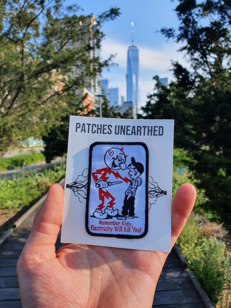 May include: A white patch with black trim featuring a cartoon illustration of a man in a red suit holding a knife and a boy in a blue shirt. The text on the patch reads "Remember Kids, Electricity Will Kill You!" The patch is held in a hand against a backdrop of a city skyline.