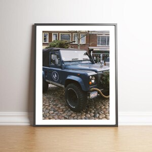 May include: A blue Land Rover Defender with a Christmas tree on top, parked on a cobblestone street in front of a brick building.