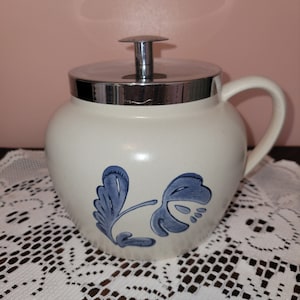 May include: A white ceramic jar with a blue floral design and a silver lid. The jar has a handle and is sitting on a white lace tablecloth.