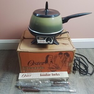 May include: An Oster electric fondue pot with a green and black finish, featuring a black handle and a temperature control dial. It sits on its original cardboard box labeled Oster Electric Fondue, accompanied by a set of six stainless steel forks.