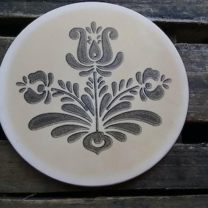 May include: A round, cream-colored ceramic plate featuring a centered, dark gray, symmetrical floral motif. The design consists of a central upright flower flanked by two smaller, outward-facing blossoms, all rendered in a stippled, textured pattern.