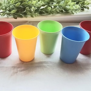 May include: Five colorful plastic cups in red, yellow, green, blue, and red are arranged on a white surface. The cups are slightly tapered and have a smooth, matte finish. Green foliage is visible in the background.